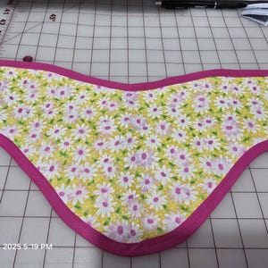 May include: A floral fabric item with a yellow background and white and purple daisy pattern, trimmed with a bright pink border. The item is likely handmade and has a curved shape.