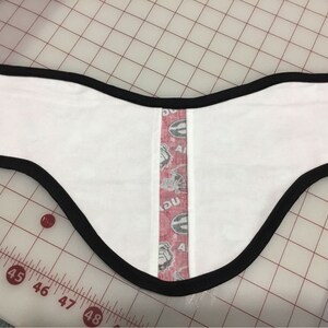 May include: White fabric neck warmer with a black trim and a red, white, and black patterned strip down the center. The fabric has a subtle texture.