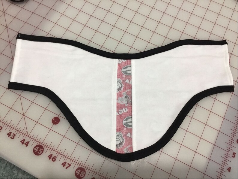May include: White fabric neck warmer with a black trim and a red, white, and black patterned strip down the center. The fabric has a subtle texture.