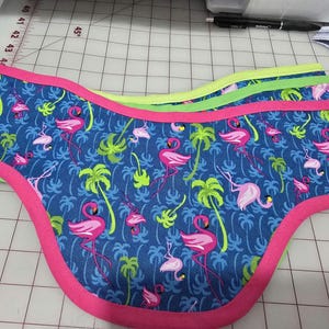 May include: A blue fabric with a pink flamingo pattern and green palm trees. The fabric has a pink trim and is folded over on itself.