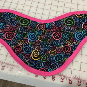 May include: A fabric item with a black background covered in colorful swirls of pink, yellow, green, and blue. The edges are trimmed with a bright pink fabric. The item is placed on a white surface with a grid pattern.