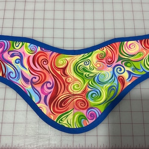 May include: A curved fabric accessory featuring a vibrant, multi-colored swirl pattern in shades of pink, orange, yellow, green, and blue. The piece is finished with a solid blue fabric binding along all edges, creating a distinct, contoured shape.