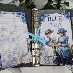 Könnte beinhalten: Ein blauer und weißer Planer mit floralem Design. Der Planer ist auf einer Seite mit dem Text "BLUE TEA" und einem Bild von zwei Frauen in blauen Kleidern, die Tee trinken, geöffnet. Der Planer hat einen Ringbinder und ist aus Filz.