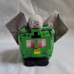 Puede incluir: Un peluche verde pixelado con alas grises y una cabeza gris. El juguete tiene ojos rojos y blancos, un pico amarillo y pies negros. El peluche está hecho de tela suave y se asemeja a un personaje de videojuego.