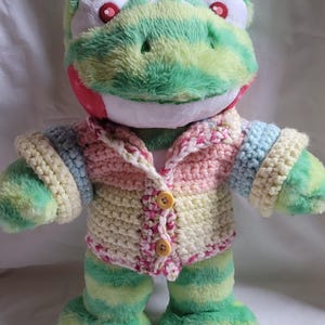 May include: A plush frog toy with a green and yellow striped body and a crocheted jacket. The jacket is cream, pink, and blue with yellow buttons. The frog has red eyes and a white mouth. The text "EXAMPLE BEAR AND OTHER ACCESSORIES NOT INCLUDED" is in the top right corner.