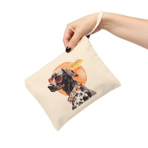 May include: A beige canvas pouch with a zipper closure. The pouch features a watercolor illustration of a black and white dog wearing sunglasses and a floral collar. The dog is in front of a yellow and orange sun.