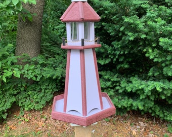 May include: A red and white wooden bird feeder shaped like a lighthouse with a white finial on top. The feeder has a clear plastic seed tray and a metal chain hanging from the top.