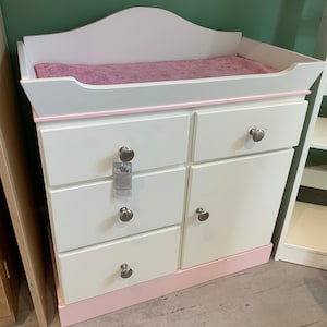 May include: White changing table with a pink trim and four drawers. The top of the changing table has a pink changing pad. The changing table has silver knobs.
