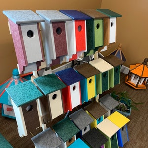 May include: A variety of birdhouses in different colors and styles, including teal, gray, brown, red, white, and green. The birdhouses are arranged on a shelf, with some stacked on top of others.