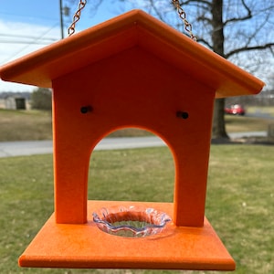 May include: Orange bird feeder with a clear glass dish and a chain for hanging. The feeder has a roof and a small opening for birds to access the food.
