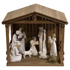 May include: A wooden nativity scene featuring carved figures in white robes, including Mary, Joseph, and the baby Jesus. The scene is set inside a rustic wooden stable with a sloped roof. Two lambs and a donkey are also present.