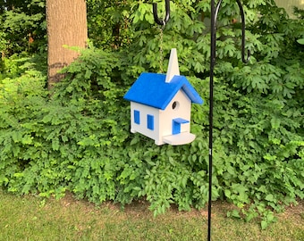 May include: A white birdhouse shaped like a church with a blue roof and blue trim. The birdhouse is hanging from a black metal hook.