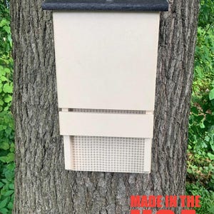May include: A white wooden bat house with a black roof and a wire mesh opening. The house is mounted on a tree trunk. The text "MADE IN THE USA" is visible in red at the bottom of the image.