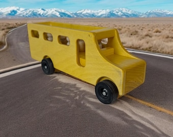 May include: A yellow wooden toy school bus with black wheels driving on a paved road. The bus is in a desert landscape with snow-capped mountains in the background.