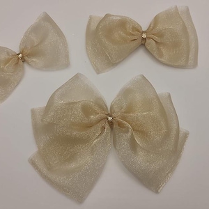 May include: Three ivory-colored organza bows with a subtle shimmer. Each bow features a delicate, layered design and a small, sparkling embellishment at the center. The bows vary in size, with the largest one prominently displayed.