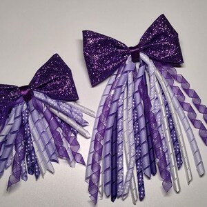 May include: Two purple glitter hair bows with white and purple curled ribbon tails.