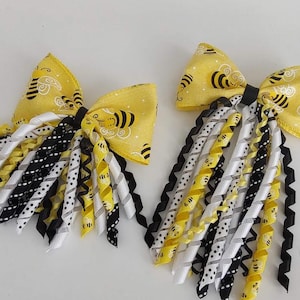 May include: Two yellow hair bows with a bee pattern. Each bow has a yellow fabric bow with black bees, and cascading ribbons in yellow, black, white, and black and white polka dots. The ribbons are curly and add a playful touch.
