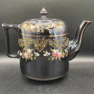 Puede incluir: Tetera de cerámica negra con tapa y asa. La tetera está decorada con motivos florales dorados y detalles en rojo, blanco y azul. El pico y el asa tienen ribetes dorados. La tetera está sobre una superficie negra.