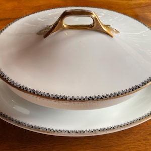 Puede incluir: Un plato de servir de porcelana blanca con una tapa con borde dorado y un borde decorativo negro y dorado. La tapa tiene un asa dorada.