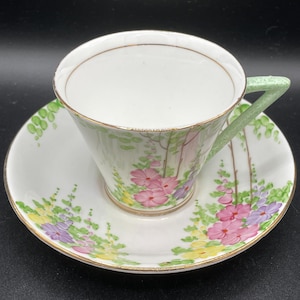 Puede incluir: Una taza de té y un platillo blancos con un patrón floral. La taza tiene un asa verde y un borde dorado. El platillo tiene un borde dorado y un patrón floral con flores rosas, amarillas y moradas.