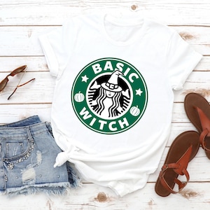 May include: A white short-sleeve t-shirt featuring a circular green graphic with the text BASIC WITCH, a central figure wearing a witch hat, and two small pumpkins. The shirt is displayed alongside blue denim shorts, brown sandals, and sunglasses.
