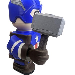 May include: A blue and brown Captain America figurine holding a gray hammer. The figurine has a blue helmet with silver accents, a white star on the chest, and white stripes on the arms. The hammer is gray with a textured handle.