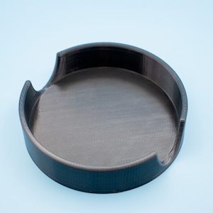May include: A black plastic round tray with a raised edge and a cutout on one side. The tray is designed to hold small items.