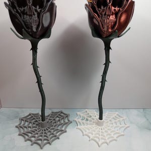 May include: Two decorative rose sculptures with skull faces. One is dark brown, the other is copper-colored. The stems are dark green with thorns, and the bases are spiderweb designs in gray and white. The roses are on a white surface.