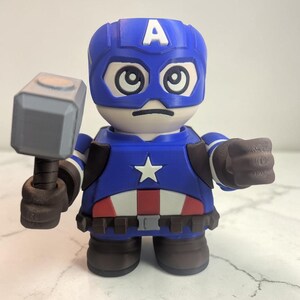 May include: A blue and white Captain America figurine holding a gray hammer. The figurine has a blue helmet with a white "A" and a white star on the chest. The figure is wearing a blue and white costume with brown gloves and boots.