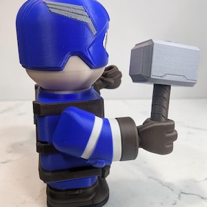 May include: A blue and white figurine of a superhero holding a gray hammer. The figurine is wearing a blue helmet with a silver emblem, a blue suit with white stripes, and brown gloves and boots. The hammer has a gray head and a brown handle.