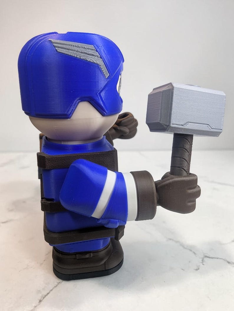 May include: A blue and white figurine of a superhero holding a gray hammer. The figurine is wearing a blue helmet with a silver emblem, a blue suit with white stripes, and brown gloves and boots. The hammer has a gray head and a brown handle.