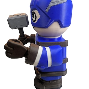 May include: A blue and white figurine of a superhero holding a gray hammer. The figurine has a blue helmet with white accents and a blue uniform with white stripes. The figurine is a toy.