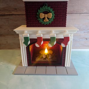 May include: A white and red miniature fireplace with a lit candle inside. The fireplace has a brick chimney with a green wreath and a gold bow. There are four red and green stockings hanging from the mantel.