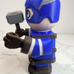 May include: A blue and white figurine of a superhero holding a gray hammer. The figurine has a blue helmet with white accents, a white face, and brown gloves and boots. The figure is wearing a blue uniform with white stripes.