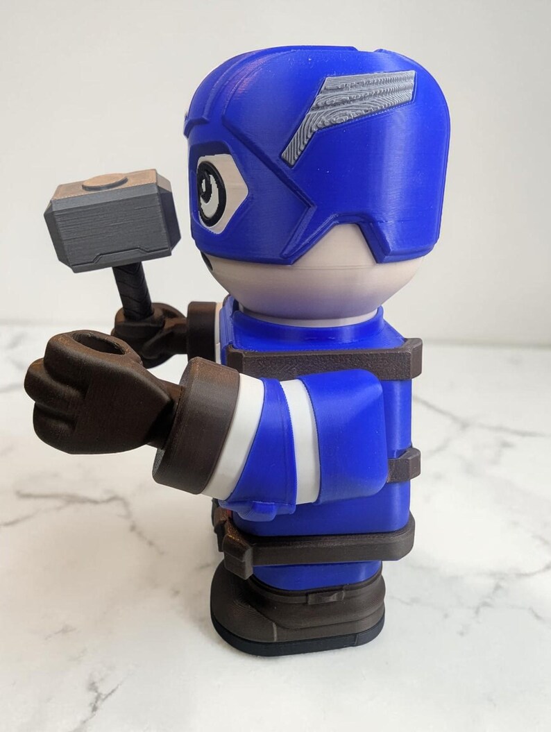 May include: A blue and white figurine of a superhero holding a gray hammer. The figurine has a blue helmet with white accents, a white face, and brown gloves and boots. The figure is wearing a blue uniform with white stripes.