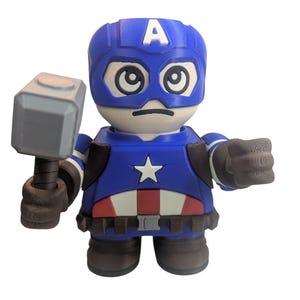 May include: A blue and red Captain America figurine holding a gray hammer. The figurine has a blue helmet with a white "A" on the forehead, a white star on the chest, and a red and white belt. The figure is in a cartoon style.