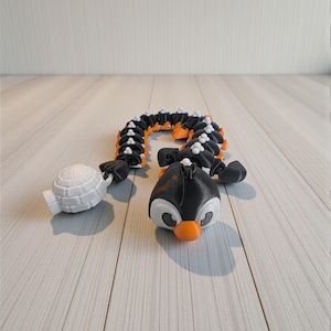 May include: A black, white, and orange articulated penguin toy with a white igloo. The penguin has a segmented body and a cute face with an orange beak. The igloo is white and has a small handle.
