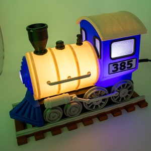 May include: A blue and yellow toy train engine with a light inside. The engine has a number "385" on the side. The train is on a wooden track.