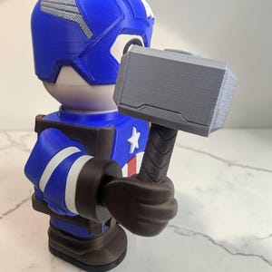 May include: A blue, white, and brown Captain America figurine holding a gray hammer. The figurine has a blue helmet with white accents, a white star on the chest, and white stripes on the arms. The hammer is gray with a brown handle.