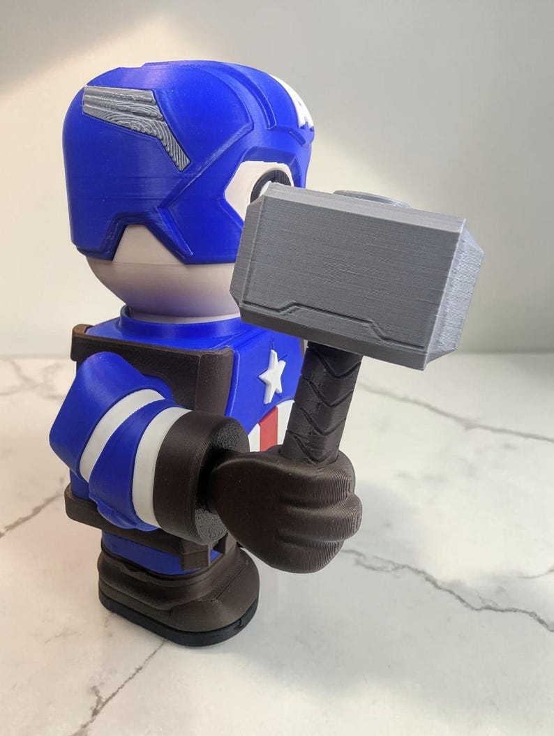 May include: A blue, white, and brown Captain America figurine holding a gray hammer. The figurine has a blue helmet with white accents, a white star on the chest, and white stripes on the arms. The hammer is gray with a brown handle.