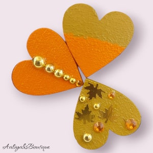 Puede incluir: Tres corazones de madera, cada uno pintado en tonos de naranja y dorado. Los corazones est&aacute;n decorados con purpurina dorada, cuentas doradas y detalles en pan de oro. Los corazones est&aacute;n dispuestos en forma de tr&eacute;bol.