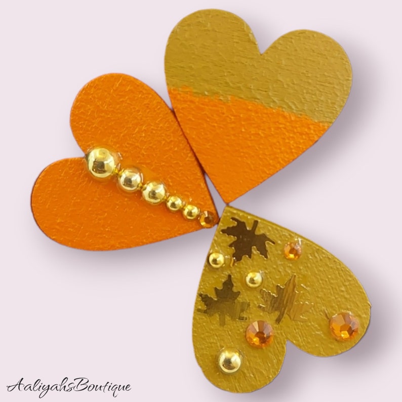 Puede incluir: Tres corazones de madera, cada uno pintado en tonos de naranja y dorado. Los corazones est&aacute;n decorados con purpurina dorada, cuentas doradas y detalles en pan de oro. Los corazones est&aacute;n dispuestos en forma de tr&eacute;bol.