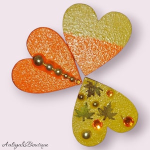 Puede incluir: Tres corazones de madera decorados con purpurina dorada y adornos. Los corazones son naranja, amarillo y una combinaci&oacute;n de naranja y amarillo. El coraz&oacute;n amarillo tiene hojas de arce de pan de oro y diamantes de imitaci&oacute;n.