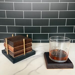 May include: A set of square dark wood coasters stacked in a gold metal holder, alongside a single matching coaster topped with a clear glass tumbler containing amber liquid and the etched word DAD in a handwritten style font.