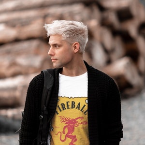 May include: A man with platinum blonde hair is wearing a white t-shirt with a Fireball Cinnamon Whisky logo. The logo is yellow with a red and orange fire design and a red demon. The man is also wearing a black cardigan and a black backpack.