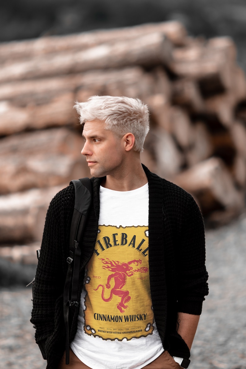 May include: A man with platinum blonde hair is wearing a white t-shirt with a Fireball Cinnamon Whisky logo. The logo is yellow with a red and orange fire design and a red demon. The man is also wearing a black cardigan and a black backpack.