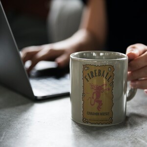 May include: A grey ceramic mug with a Fireball Cinnamon Whisky logo. The logo features a red demon with a yellow background and the text "Fireball Cinnamon Whisky".