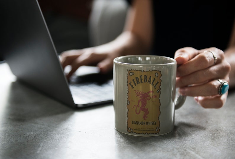 May include: A grey ceramic mug with a Fireball Cinnamon Whisky logo. The logo features a red demon with a yellow background and the text "Fireball Cinnamon Whisky".