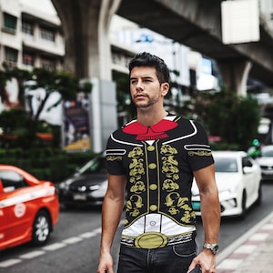 Puede incluir: Un hombre con una camiseta negra con un diseño dorado y blanco de un atuendo tradicional mexicano. La camiseta tiene una corbata de lazo roja y un cinturón dorado. El hombre lleva pantalones vaqueros negros y lleva una bolsa negra.