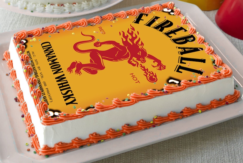 May include: A white cake with orange frosting and sprinkles. The cake is decorated with a printed image of a Fireball Cinnamon Whisky bottle label. The label features a red demon with flames and the text "Fireball", "Red", "Hot", and "Cinnamon Whisky".